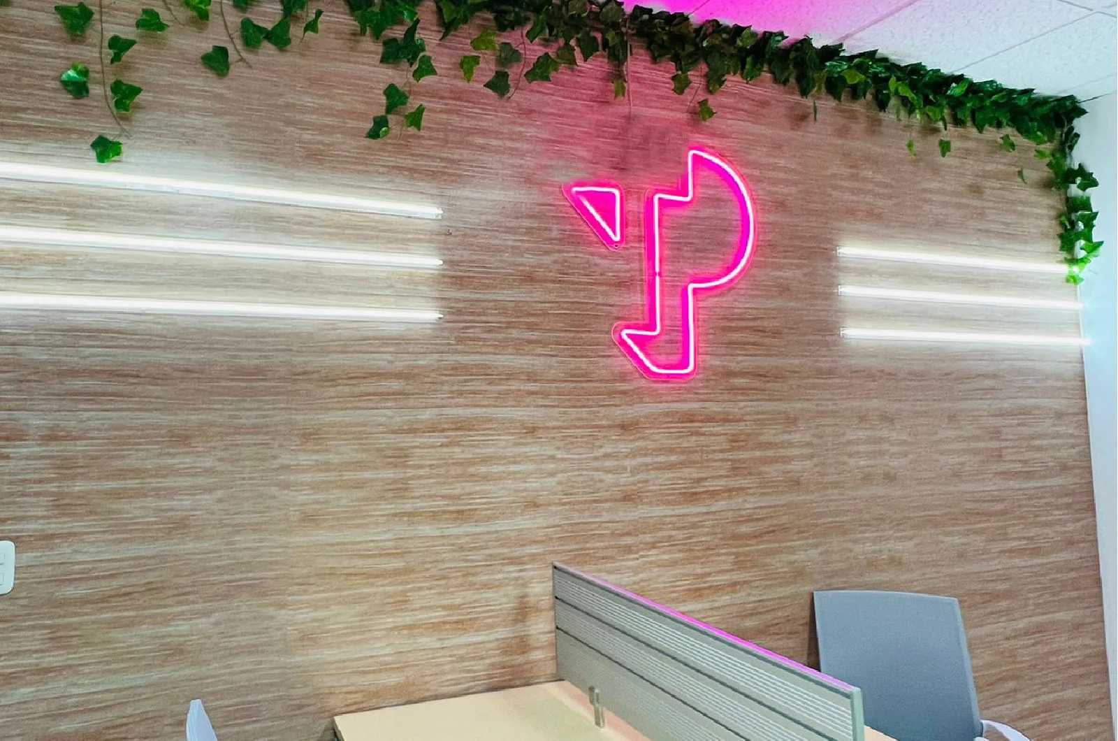TP logo in pink neon on a wooden wall with greenery and office desk in the foreground.