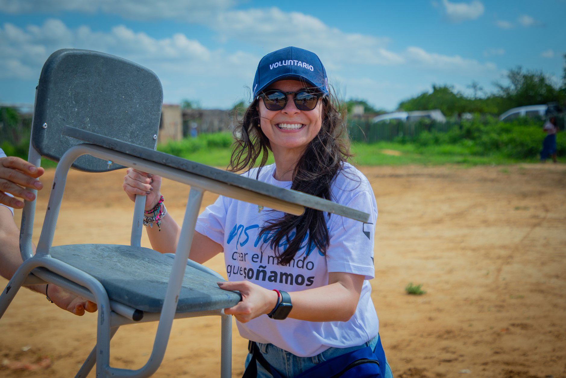 A smiling person wearing a "voluntario" hat holds a chair outdoors. 