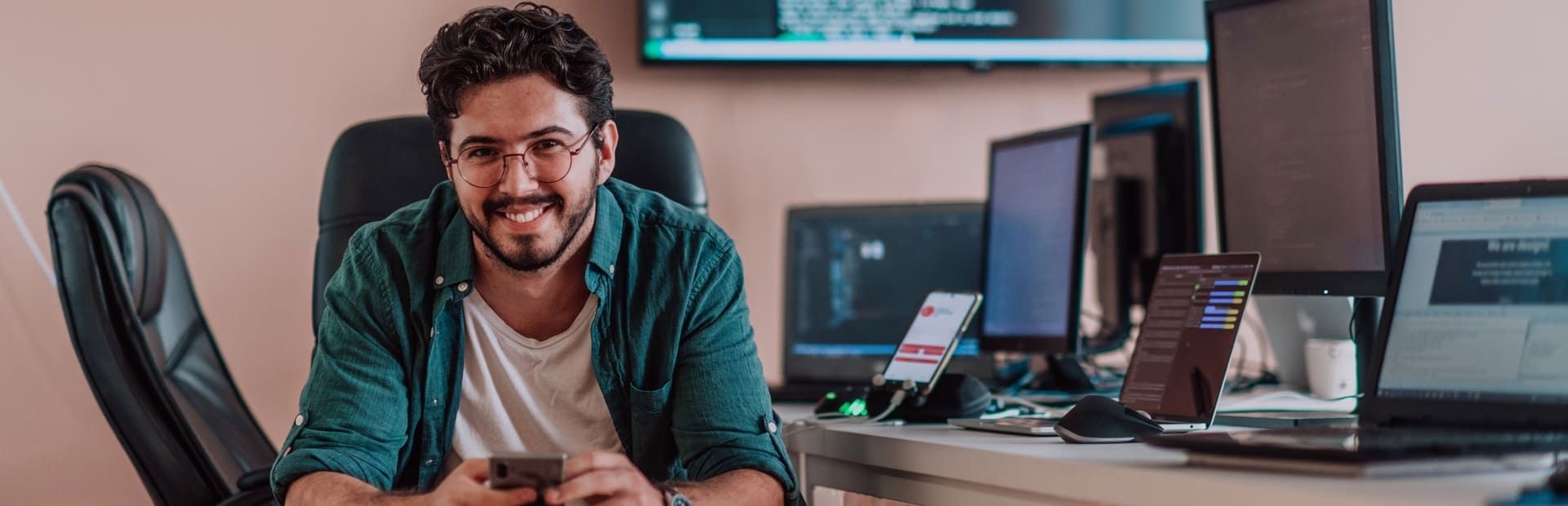 Man smiling at his desk in a tech workspace with multiple monitors and devices, reflecting TP’s digital focus.