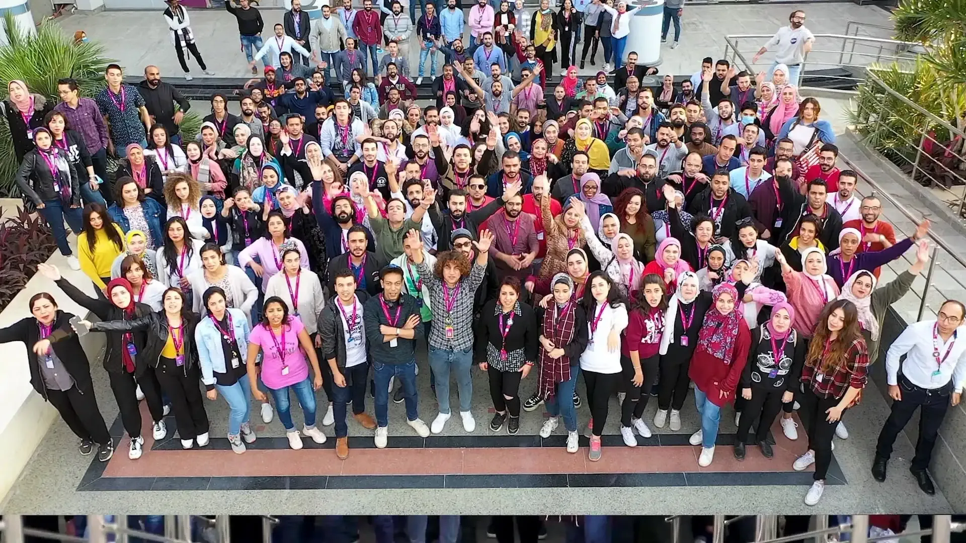Large group of people gathered outdoors, many wearing colorful clothing and smiling while waving at the camera. Large group of people gathered outdoors, many wearing colorful clothing and smiling while waving at the camera.