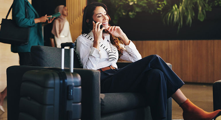 Businesswoman in modern airport lounge talking on phone with suitcase, dressed in smart casual attire.