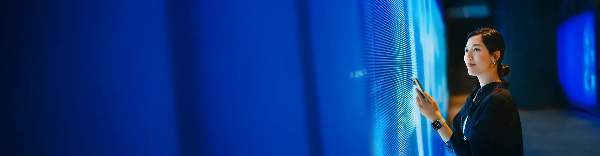 Asian woman holding a smartphone in front of a modern digital display.