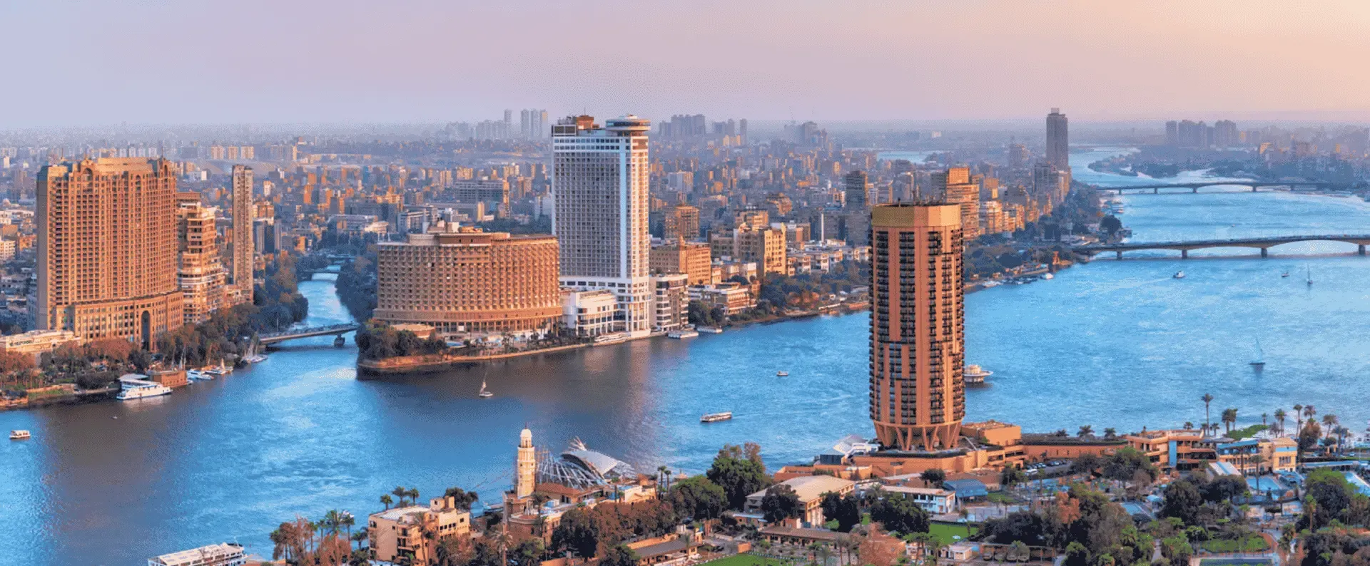 Cairo skyline with skyscrapers along the Nile River, bridges and boats under a warm sunset sky.