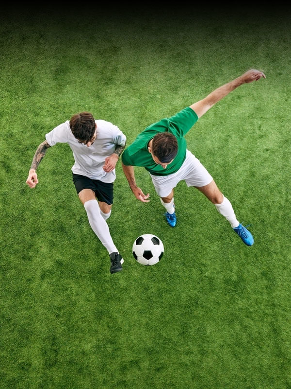 two soccer players, one in white and one in green, jump to head a ball on a grassy field
