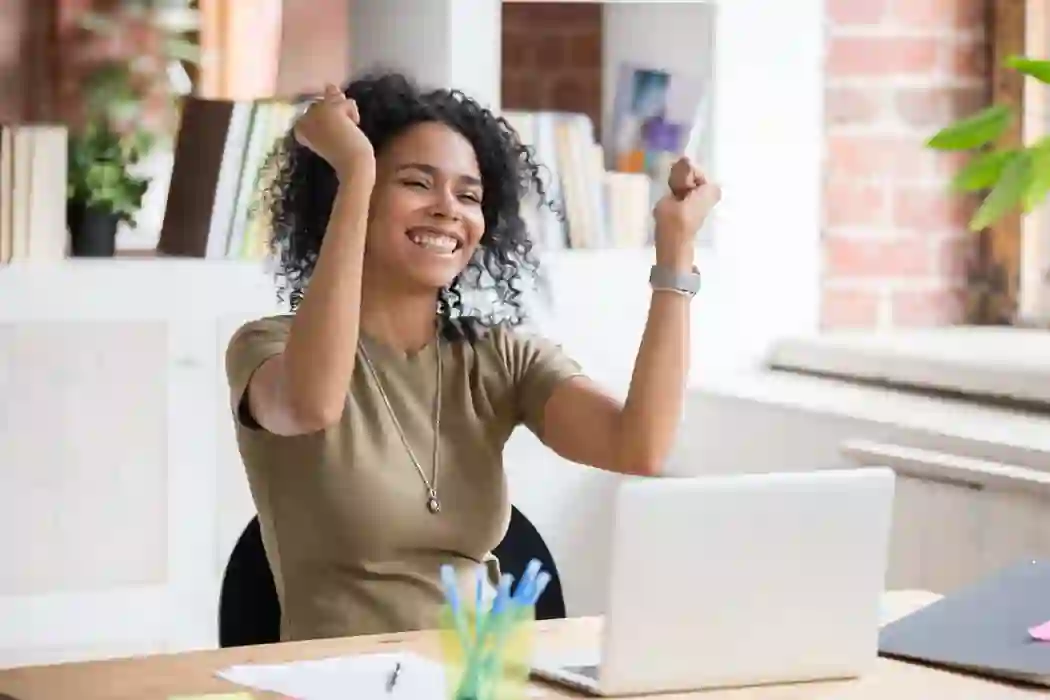 Afro-descendant woman celebrating with raised arms while working on her computer in a white office with rustic brick walls.