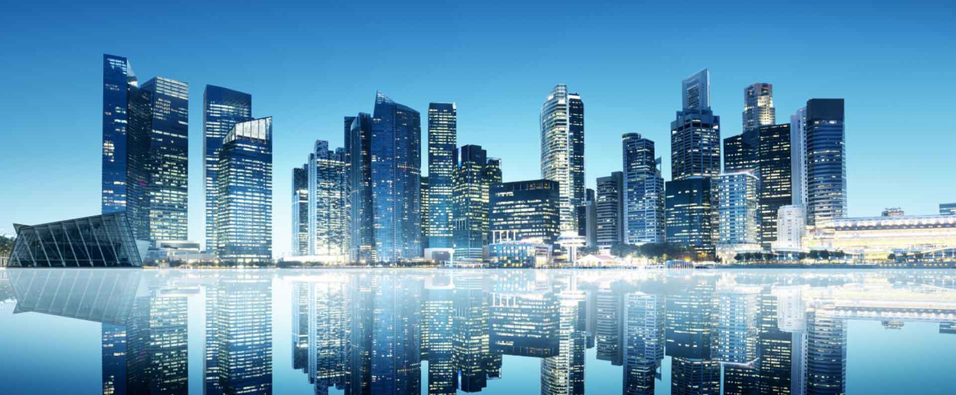 Skyscrapers stand tall, reflecting in a calm urban waterfront at dusk, with city lights glowing against a clear blue sky.