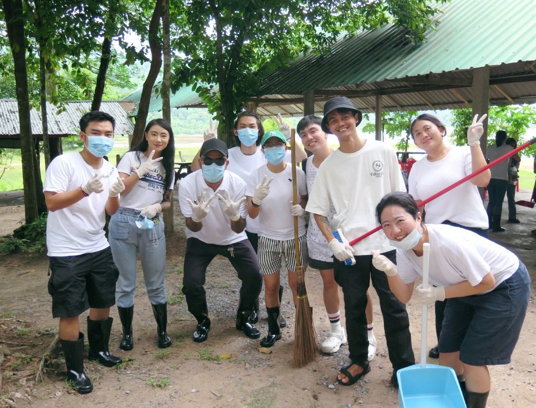 A cheerful team of volunteers in white shirts poses with cleaning tools, ready for a community cleanup in a lush, outdoor setting.
