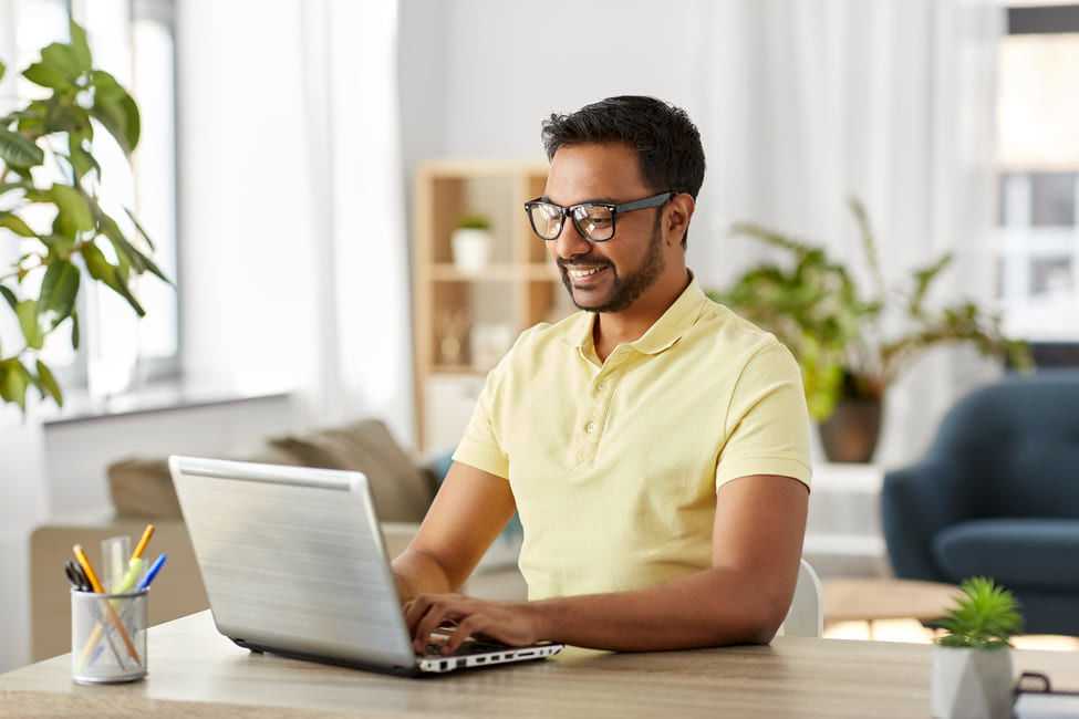 Man wearing glasses and a yellow shirt working on his laptop in a bright, plant-filled home.