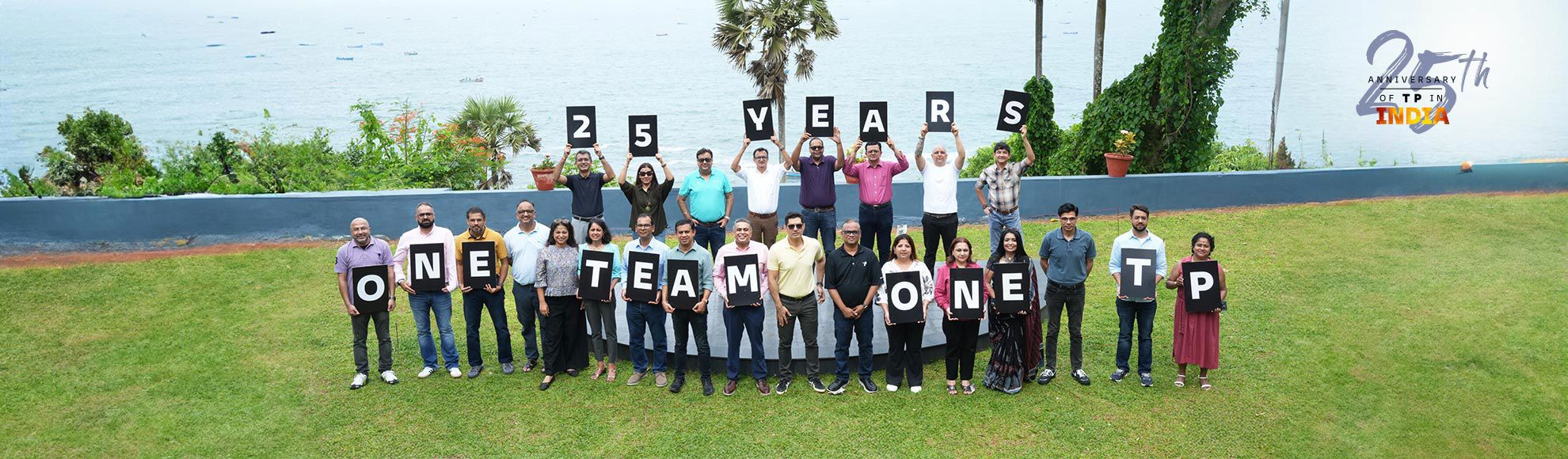 Team members celebrating TP's 25th anniversary by holding letter boards spelling '25 Years One Team One TP' at an outdoor waterfront event.