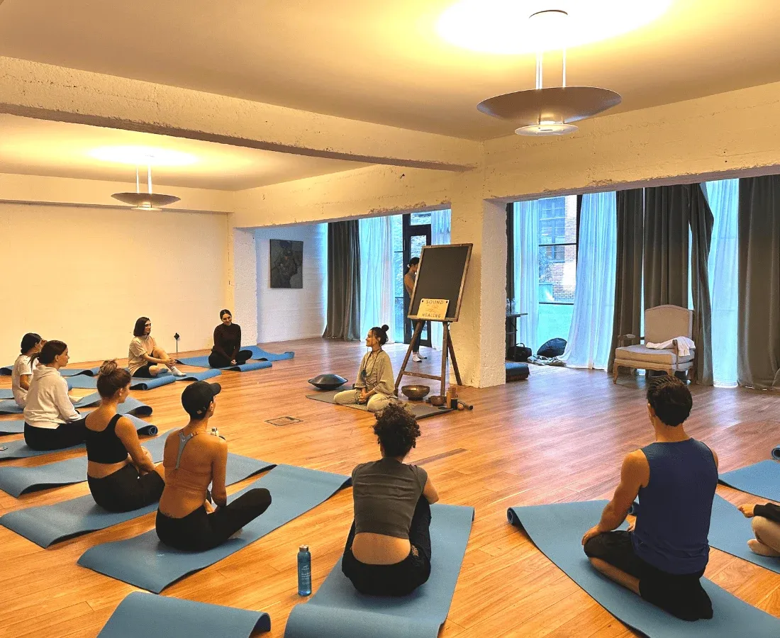 Participants sit on yoga mats, facing an instructor by an easel labeled "sound healing''.