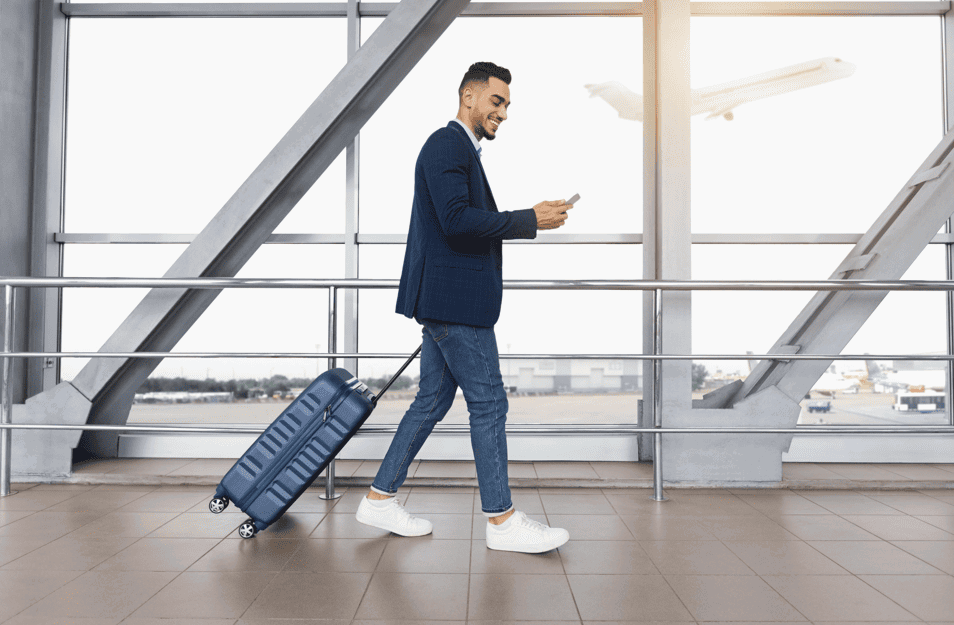 a man in casual attire pulls a suitcase while checking his phone, walking through an airport terminal with large windows and a plane taking off outside.