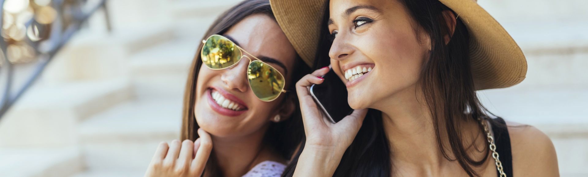Two women smiling and posing together outdoors, one holding a phone and the other wearing sunglasses and a hat.