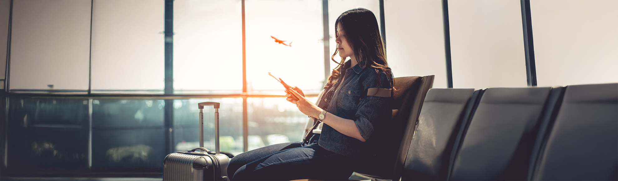 Traveler sitting in an airport lounge using a smartphone with luggage beside them and an airplane taking off in the background.