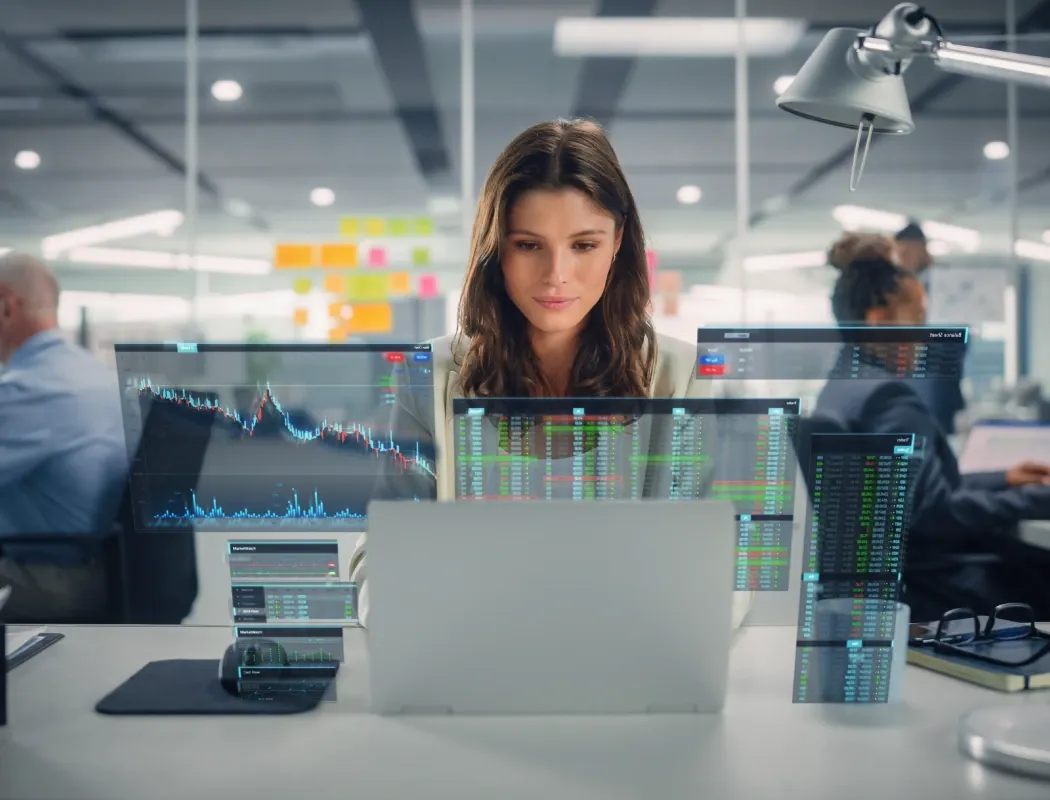 A professional woman focuses intently on analyzing real-time financial data and stock market trends on multiple transparent holographic screens in a modern, high-tech office workspace.