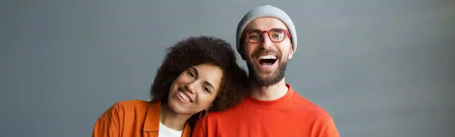 Two smiling people posing together, one leaning on the other’s shoulder against a gray background.