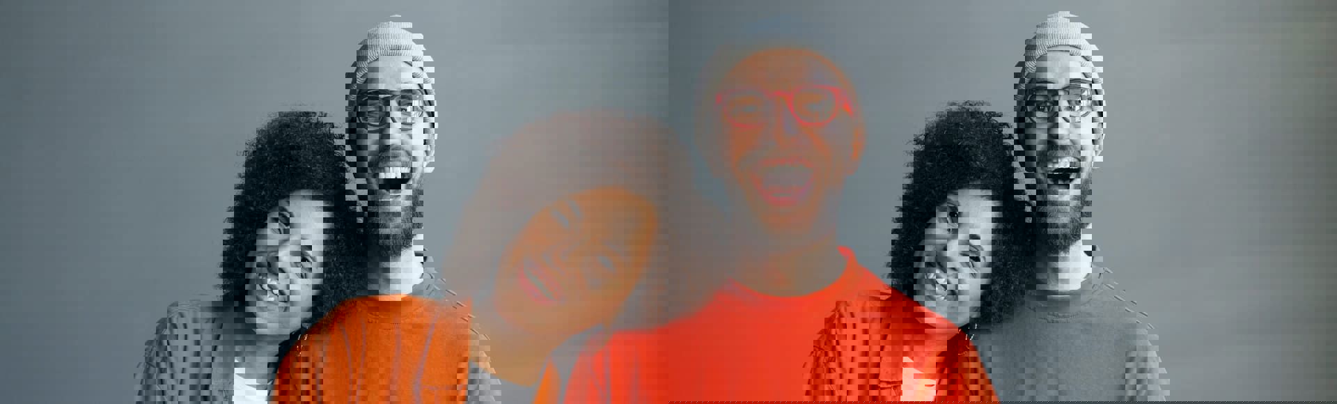 Two smiling people posing together, one leaning on the other’s shoulder against a gray background.