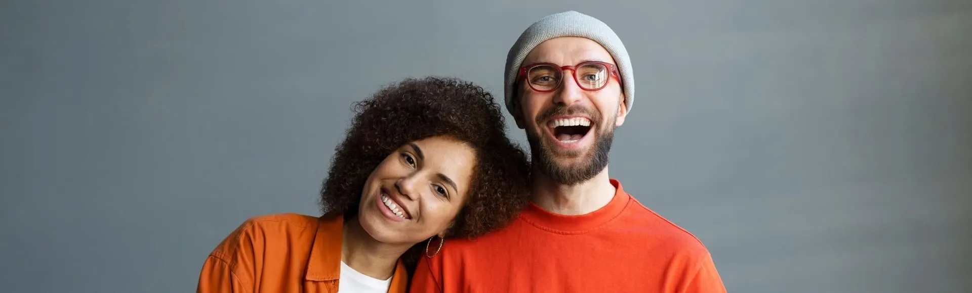 Two smiling people posing together, one leaning on the other’s shoulder against a gray background.