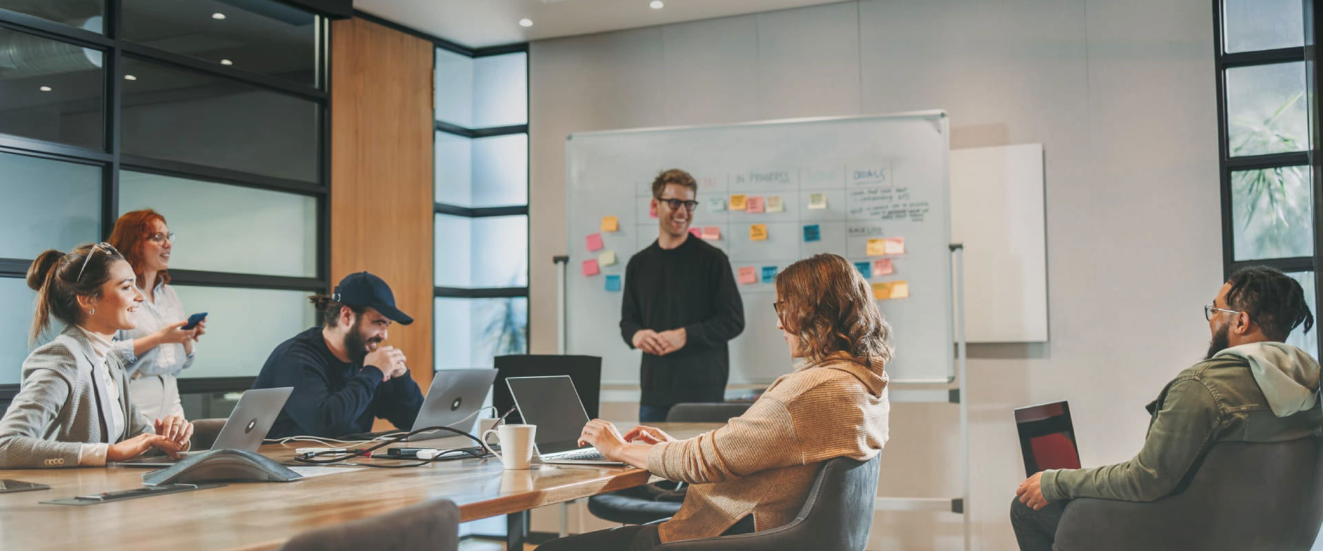 Group of people in a modern meeting room with laptops and sticky notes on the wall, one person standing and presenting.