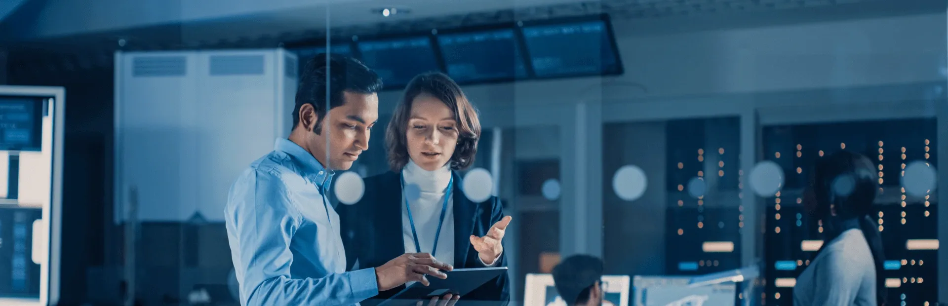 Two professionals reviewing information on a tablet inside a technology operations room.