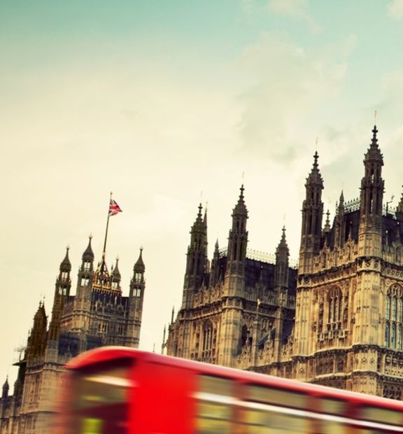 A red local bus drives past Buckingham Palace in a classic London city scene.