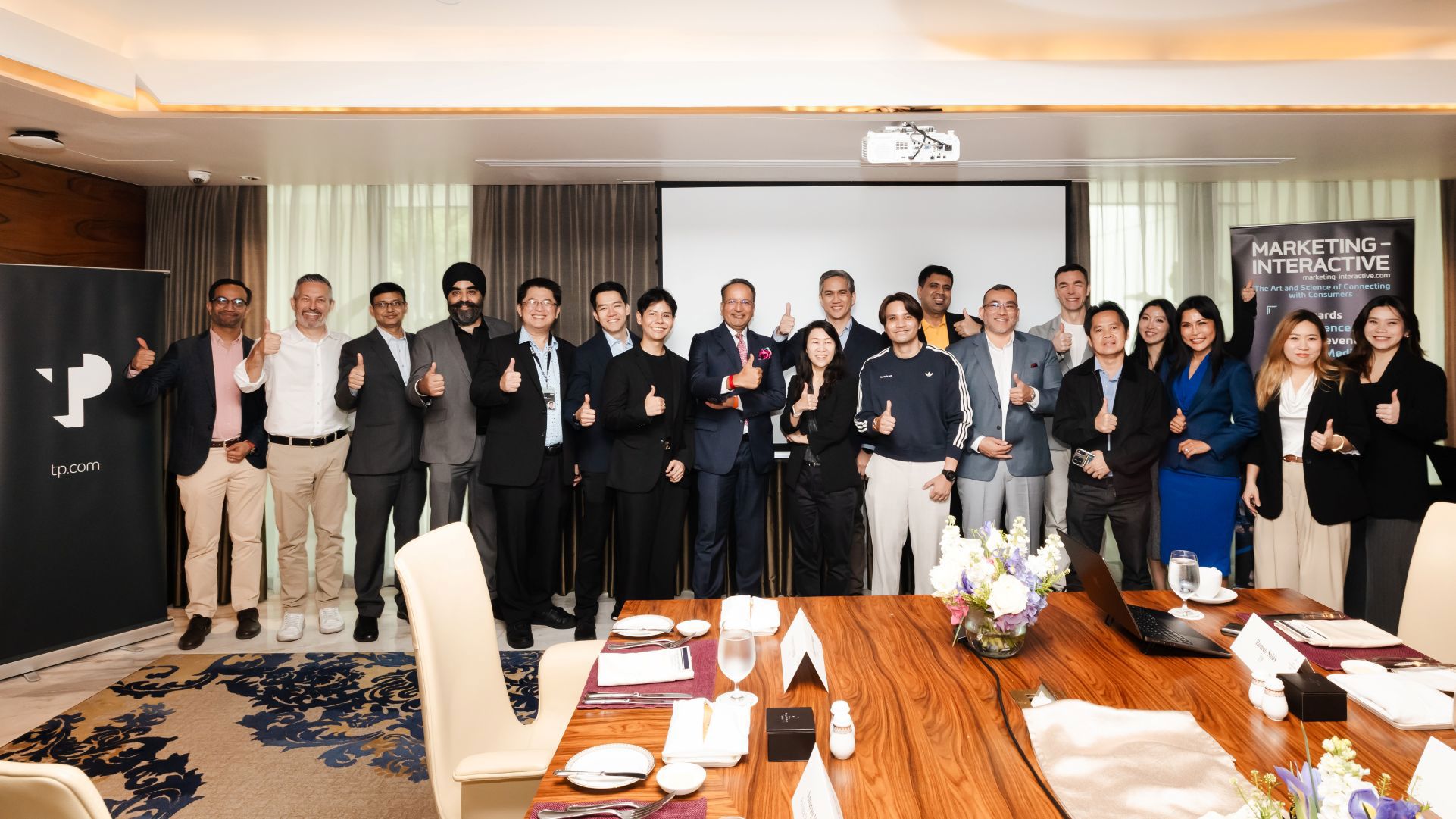 A diverse group of business professionals poses with thumbs-up gestures in a conference room, flanked by "tp" and "Marketing Interactive" banners, celebrating a successful collaboration or achievement. 