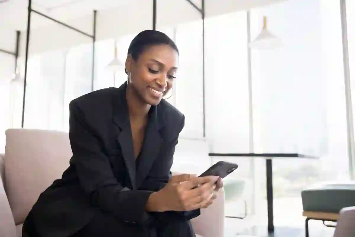 Smiling African business woman browsing her cellphone on a modern glasswalls poffice