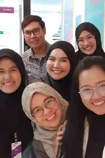 Six people smiling at the camera in a brightly lit office, some wearing hijabs and others glasses.