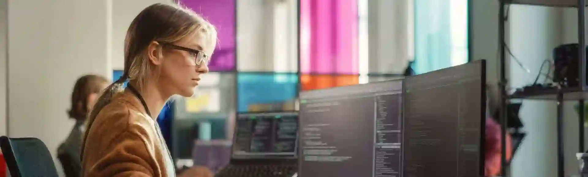 Blonde woman wearing glasses focused on coding across dual monitors, illustrating the delivery of AI data services.
