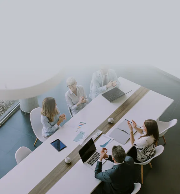 A team of business professionals claps together around a conference table, celebrating a successful meeting or milestone in a bright modern office. 