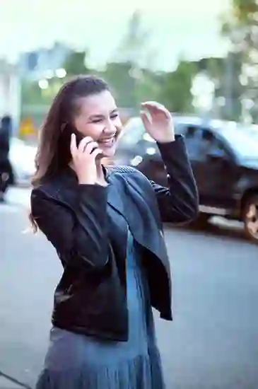 A person is happily talking on a phone, smiling, and gesturing with a hand. they are standing on a street with blurred cars and trees in the background.