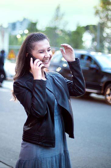 A person is happily talking on a phone, smiling, and gesturing with a hand. they are standing on a street with blurred cars and trees in the background. A person is happily talking on a phone, smiling, and gesturing with a hand. they are standing on a street with blurred cars and trees in the background.