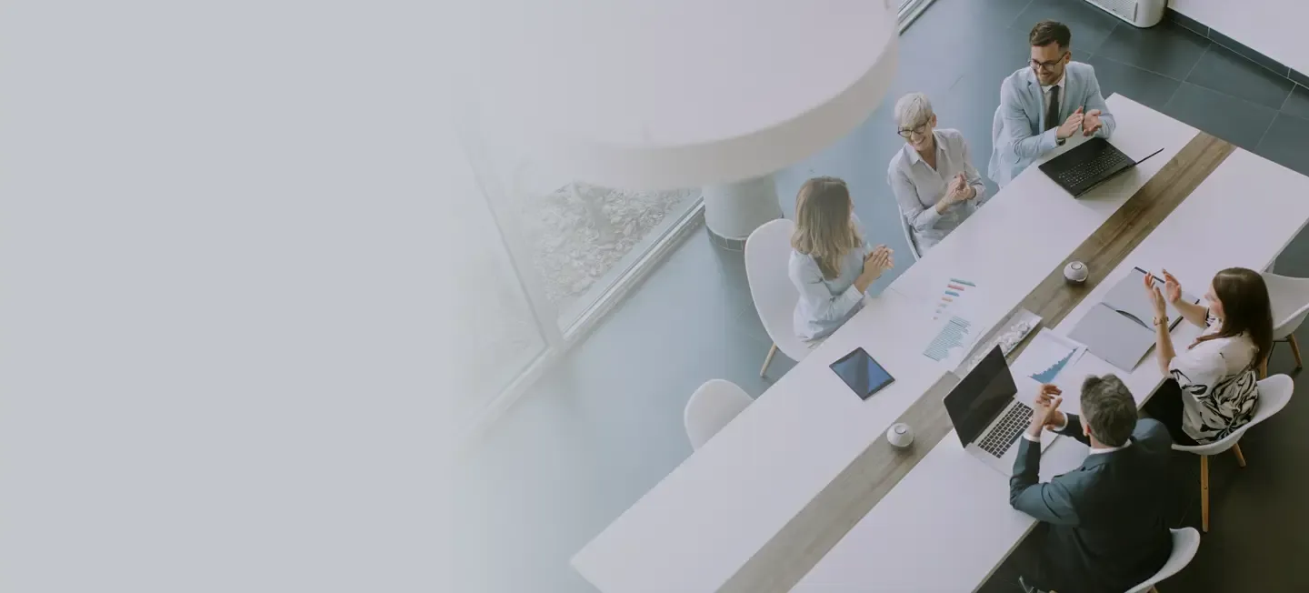 A team of business professionals claps together around a conference table, celebrating a successful meeting or milestone in a bright modern office. 