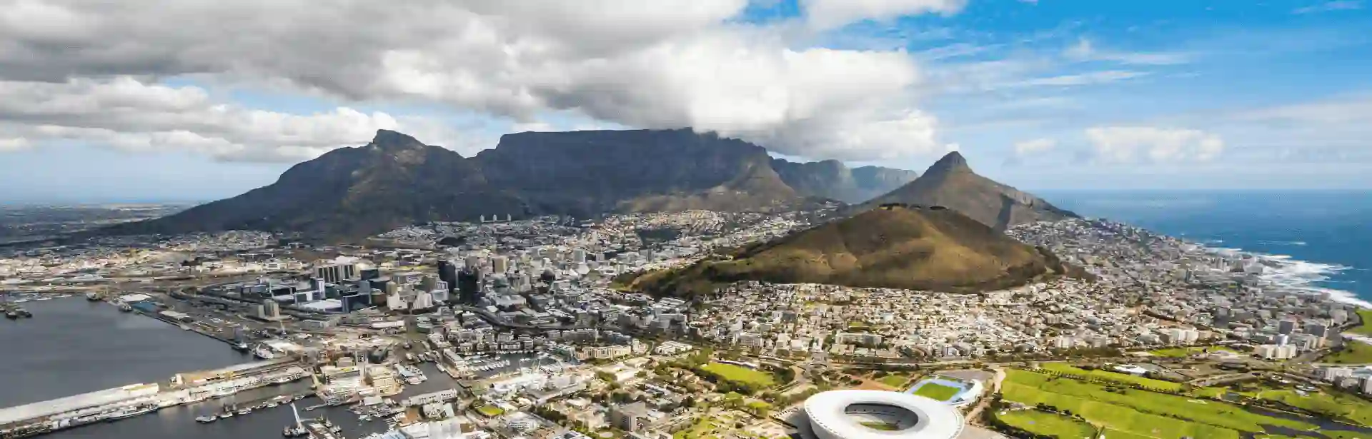 Coastal view of Cape Town with Table Mountain, urban buildings, harbor and stadium under cloudy sky.