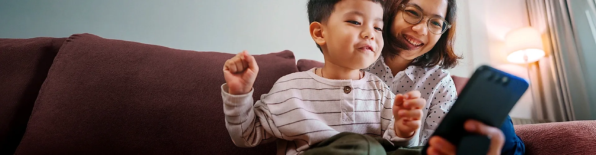 Mother and child smiling while using a smartphone together on a sofa.