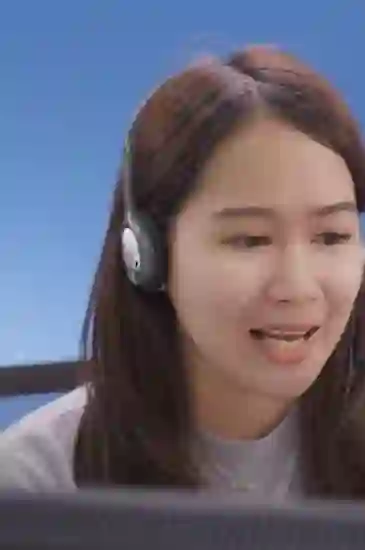 A woman wearing a headset speaks into a microphone, seated in front of a computer screen, with a plain blue background behind her.