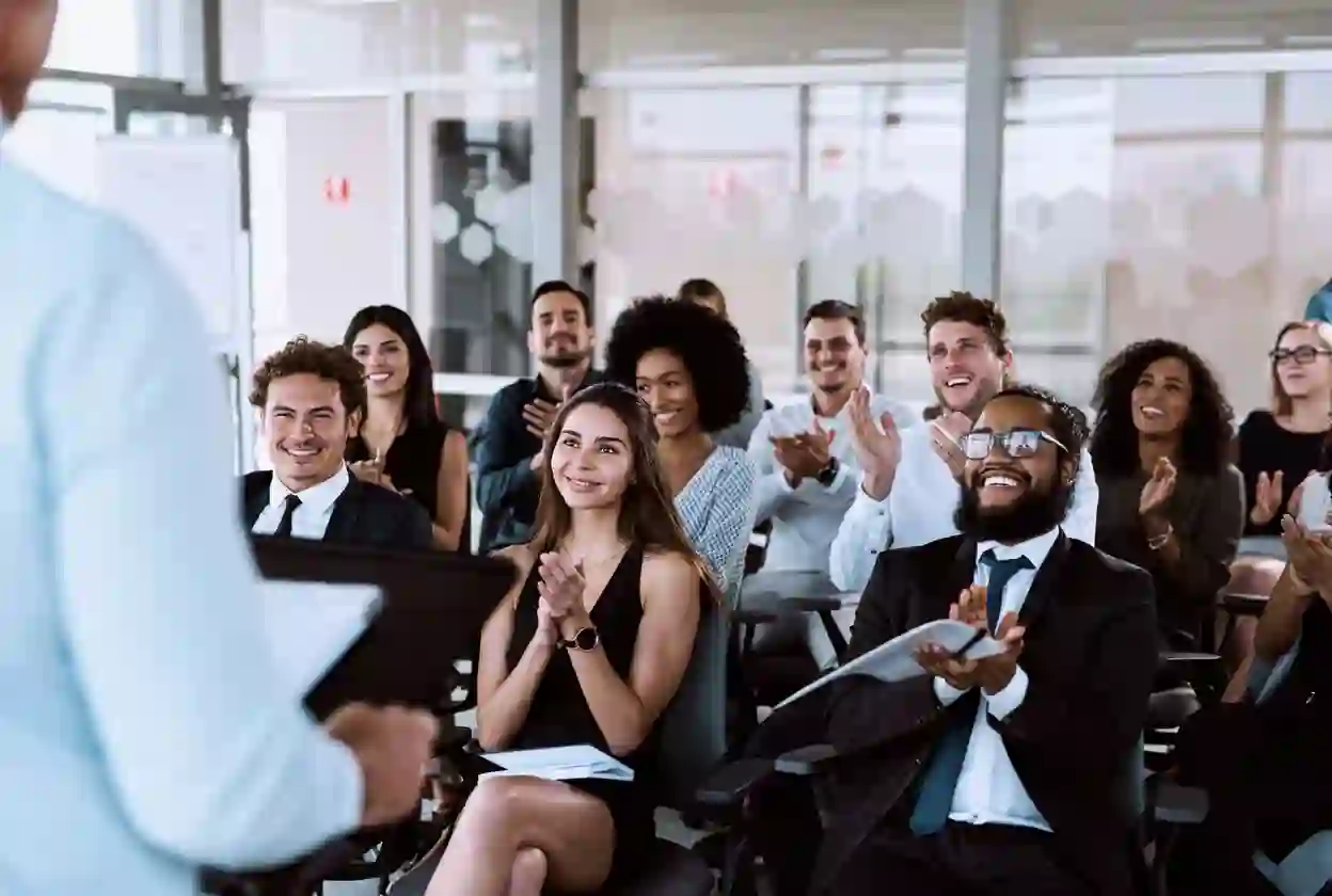 Group of diverse professionals sitting in chairs clapping and smiling during a business presentation
