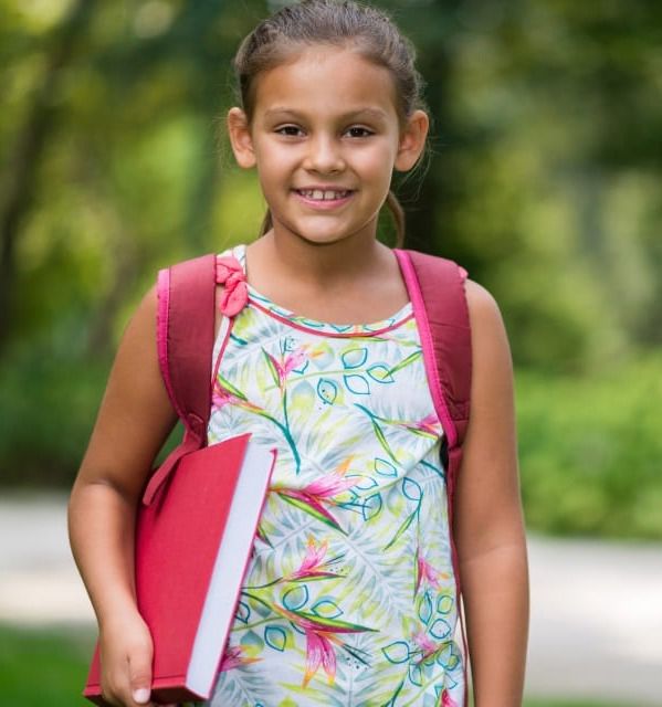 Young girl smiling while holding a red book and wearing a backpack.