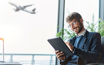 Business traveler sitting in an airport lounge using a digital tablet with a blurred airplane taking off in the background.
