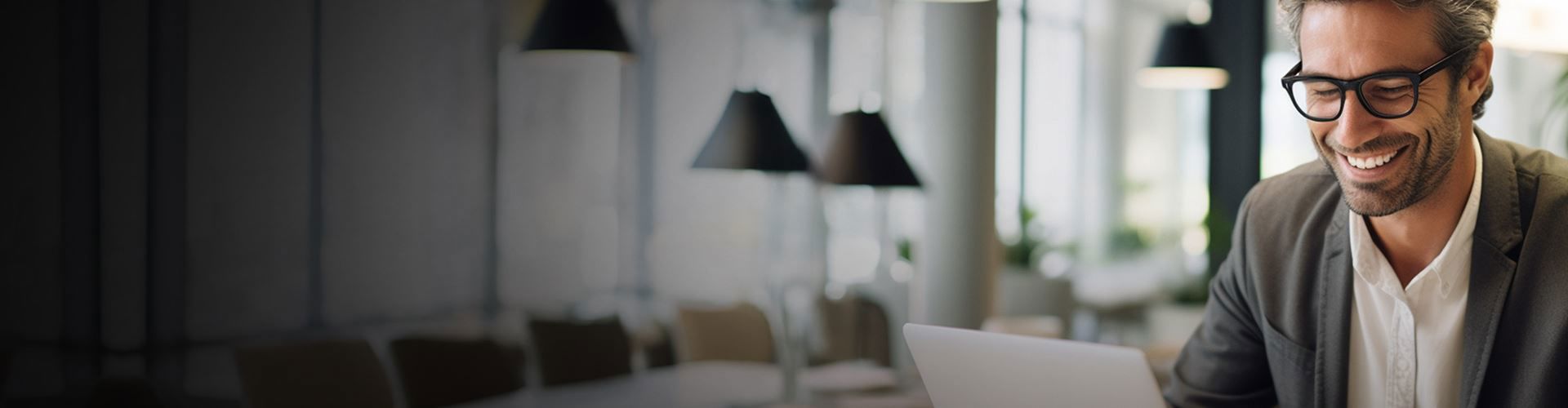 a person wearing glasses and a suit smiles while using a laptop in a well-lit, modern office area with several hanging lamps and blurred background elements.