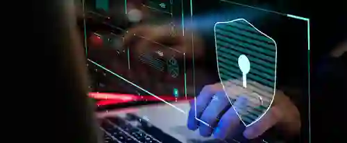 hands type on a laptop keyboard with a digital overlay showing a shield and padlock, indicating cybersecurity, set in a dimly lit environment with colorful electronic displays.