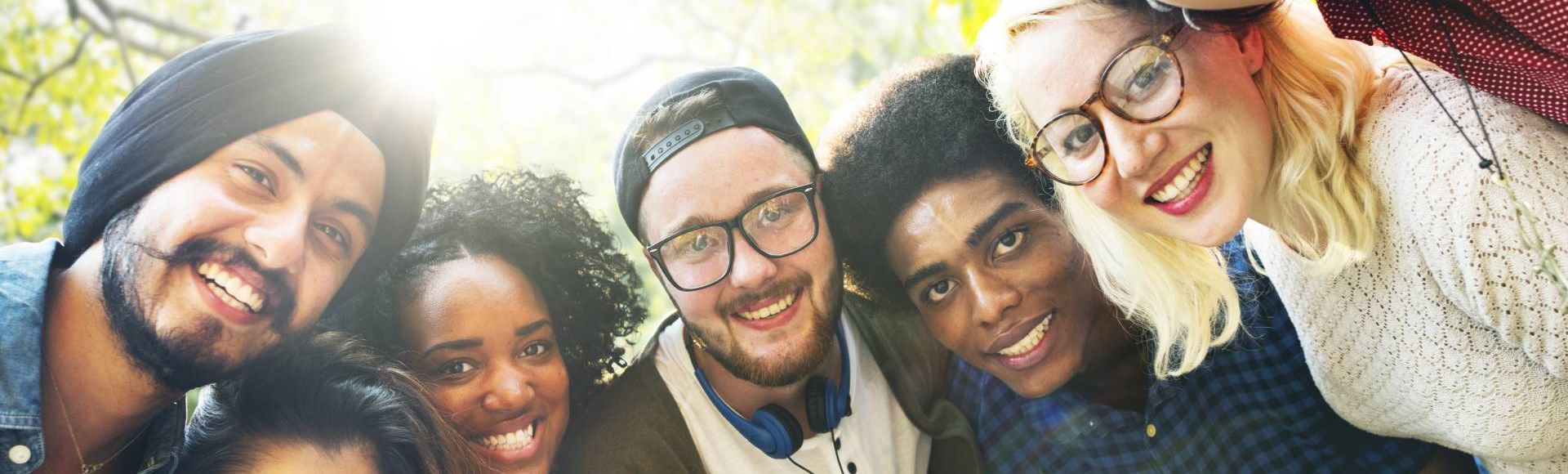 A diverse group of smiling people gathered outdoors in a sunlit, tree-filled setting.