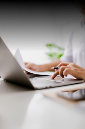 Person working on a laptop while holding a pen and papers 