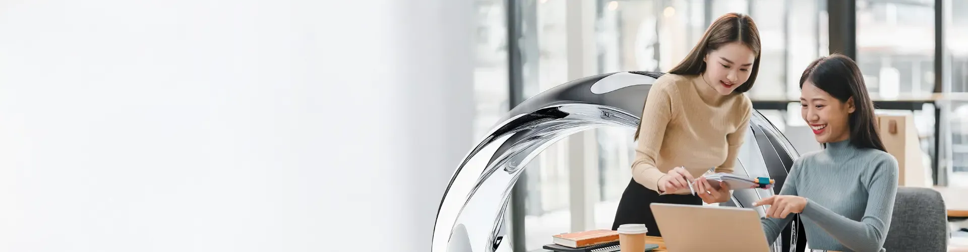 Two professional women collaborate happily at work, discussing ideas on a laptop in a bright modern office.