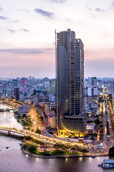 A tall skyscraper stands illuminated against a twilight sky, surrounded by urban cityscape and intersecting roads with bustling traffic.