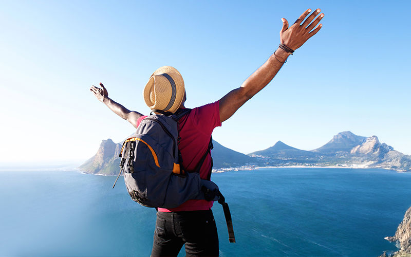 Traveler with arms raised enjoying panoramic view of blue ocean and rugged mountains from a high cliff, wearing a straw hat and backpack under clear sunny sky.