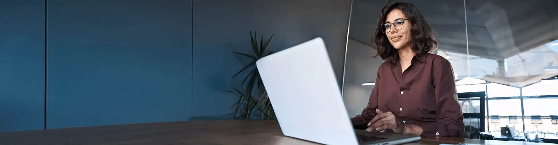 Businesswoman wearing glasses and a brown blouse working on her laptop in a modern office with glass walls.