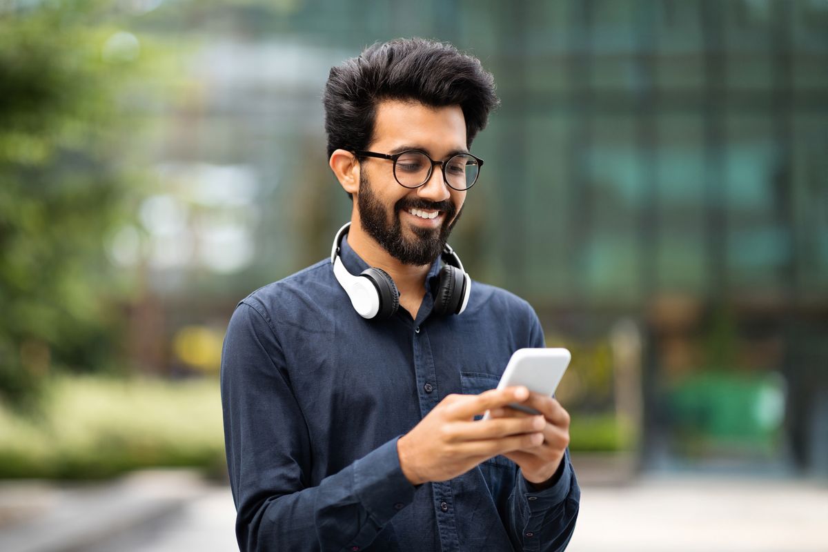 A person with headphones around their neck is smiling while looking at a smartphone outdoors, with a blurred background of greenery and possibly buildings.