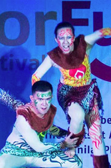Two male dancers in vibrant tribal costumes perform on a stage with blue lighting. Two male dancers in vibrant tribal costumes perform on a stage with blue lighting.