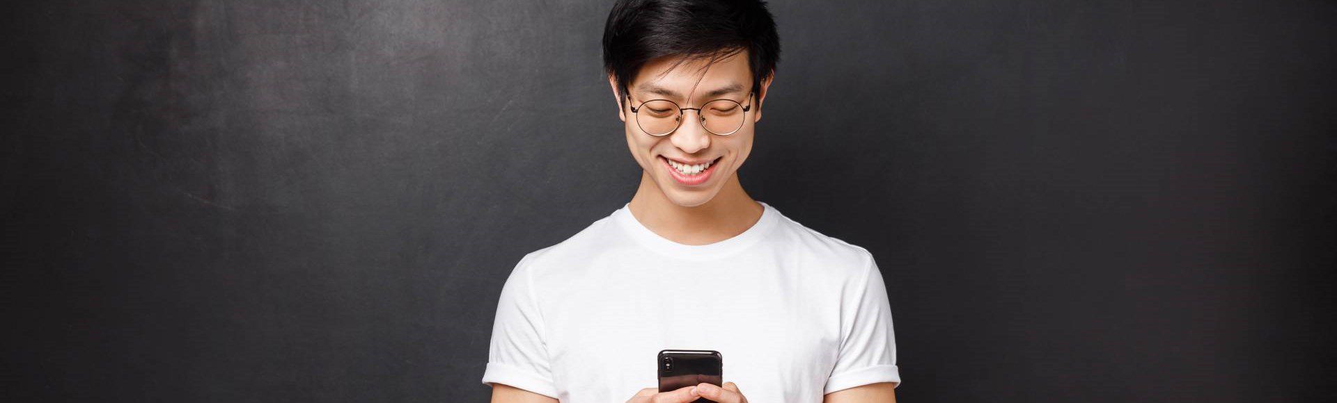 Smiling young Asian man happily reading good news on his smartphone.