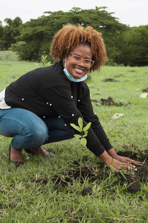 a person crouches while planting a small sapling in a grassy field, smiling and wearing a black jacket and a face mask under their chin; trees are visible in the background. a person crouches while planting a small sapling in a grassy field, smiling and wearing a black jacket and a face mask under their chin; trees are visible in the background.